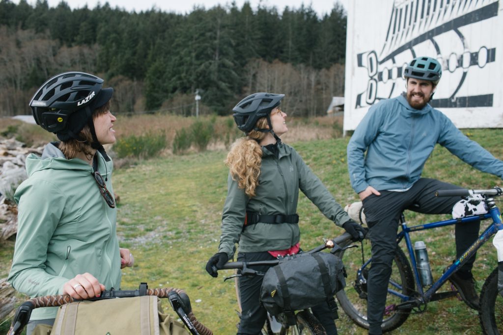 Three people on bicycles infront of a long house on Cormorant Island